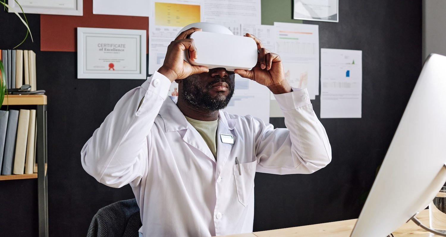 African male doctor in white coat sitting at his workplace with virtual reality goggles playing video game
