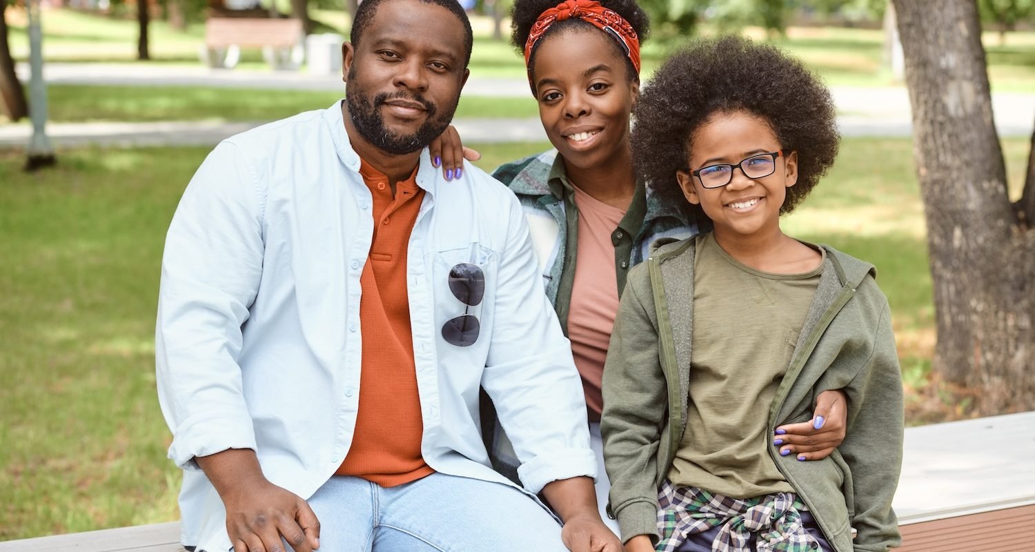 Cheerful African family sitting on bench in park on summer day
